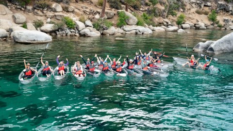 Group of kayakers in clear kayaks on a turquoise lake, near rocky shoreline.