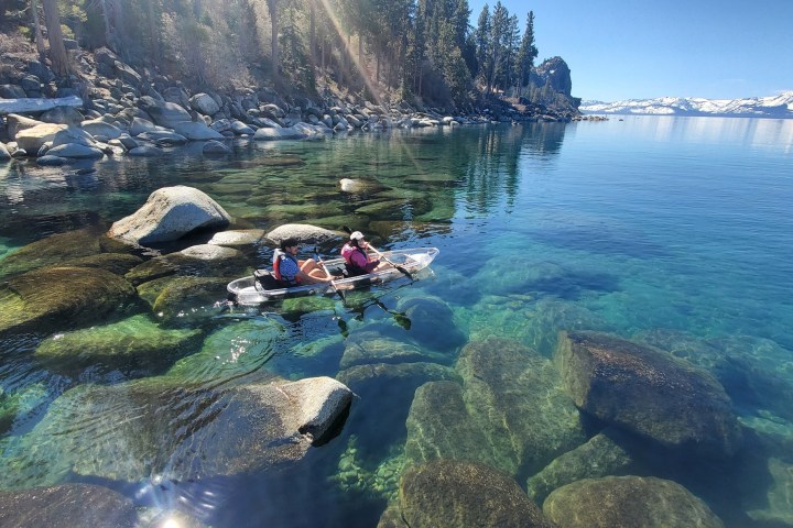 Two people kayaking on a clear lake with rocks visible underwater, surrounded by trees and distant mountains.
