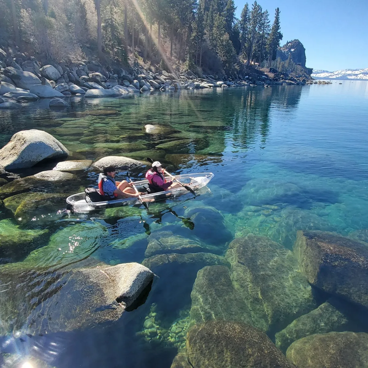 Two people kayaking on a clear lake with rocks visible underwater, surrounded by trees and distant mountains.