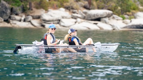Two people in a transparent kayak on a lake near rocky shore.