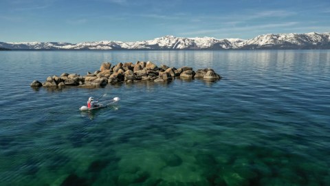 Kayaker paddling near rocky formation on clear lake with snowy mountains in background.