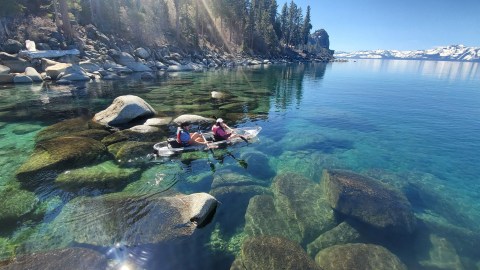 Two people kayak in a clear lake near rocky shore and pine trees, with snow-capped mountains in the background.