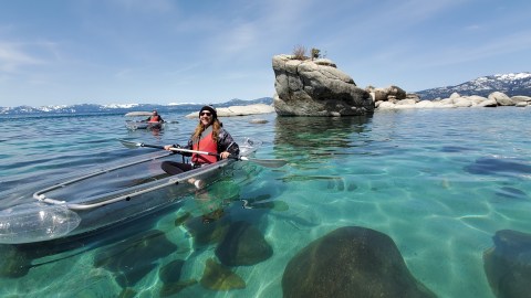 Two people kayaking in clear kayaks on a turquoise lake near large rocks and mountainous background.
