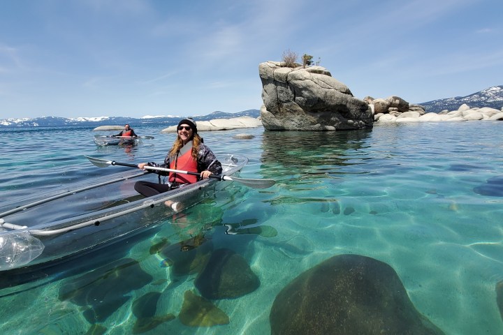 Two people kayaking in clear kayaks on a turquoise lake near large rocks and mountainous background.