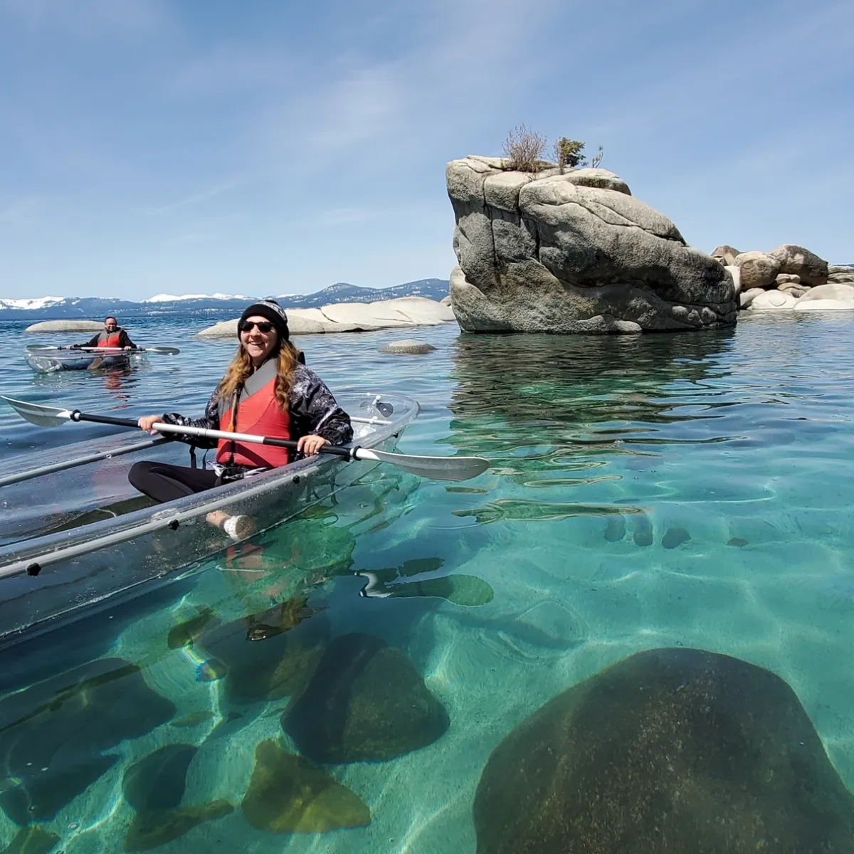 Two people kayaking in clear kayaks on a turquoise lake near large rocks and mountainous background.