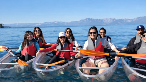 Group kayaking in clear canoes on a lake with mountains in the background.