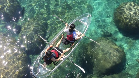 Two people kayak in clear water, revealing rocks beneath the surface.