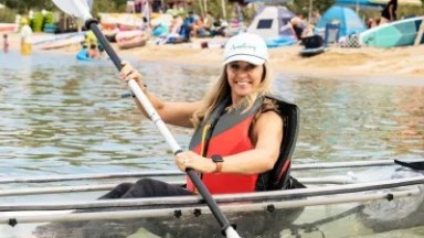 Smiling woman in hat kayaking on clear lake near busy beach with colorful umbrellas.