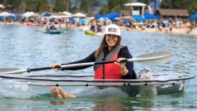 Woman kayaking on clear waters, wearing a cap and life jacket, with a beach in the background.