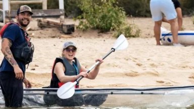 Two people with a transparent kayak near a sandy shore, one paddling and the other smiling.