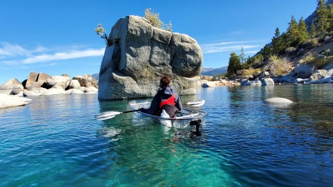 Person kayaking in clear water near large rocks and trees under a blue sky.