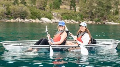 Two women kayaking in a clear kayak on a lake surrounded by trees.