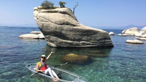Person in a clear kayak pointing at a large rock formation in clear blue water.