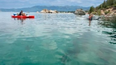People kayaking on a clear turquoise lake near rocky shore under a cloudy sky.