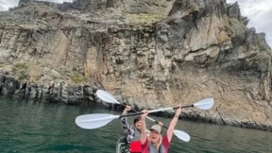 Two people kayaking near a rocky cliff under a cloudy sky.