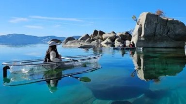 Person kayaking in a clear kayak on a crystal-clear lake near large rocks.
