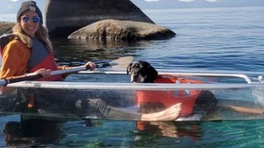 Woman and dog in a clear kayak on a calm lake with rocks and mountains in the background.