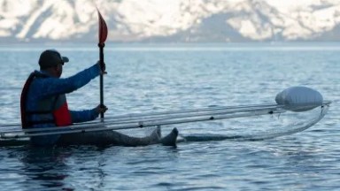 Person kayaking in clear kayak on lake with snowy mountains in background.