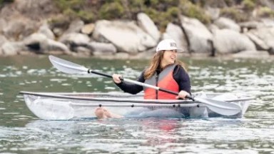Woman kayaking on clear water in a transparent kayak, wearing a red life jacket.