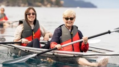 Two people kayaking on a lake, wearing life jackets and sunglasses, with a forested shore in the background.
