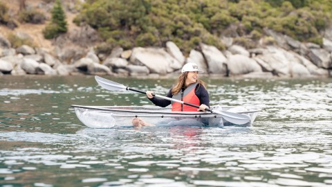 Woman kayaking in a clear kayak on a calm lake near rocky shoreline.