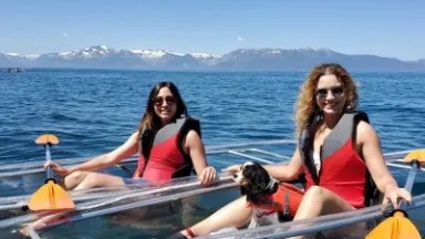 Two women in red life vests kayaking on clear water with mountains in the background.