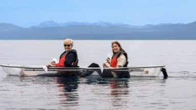 Two people in a transparent kayak on a calm lake with mountains in the background.